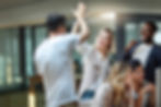 Two colleagues high-fiving in the office, while other colleagues are celebrating