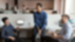 A group of colleagues laughing together while sitting around a laptop in a casual office environment.